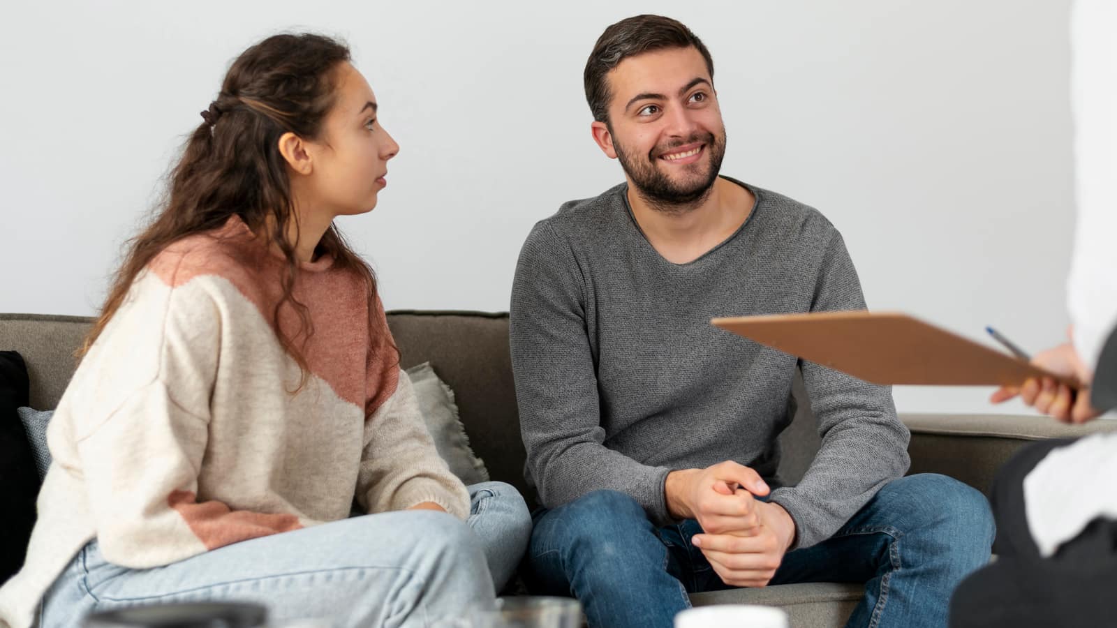 A young couple sitting together on a sofa in a therapy session, turning toward a therapist holding a clipboard