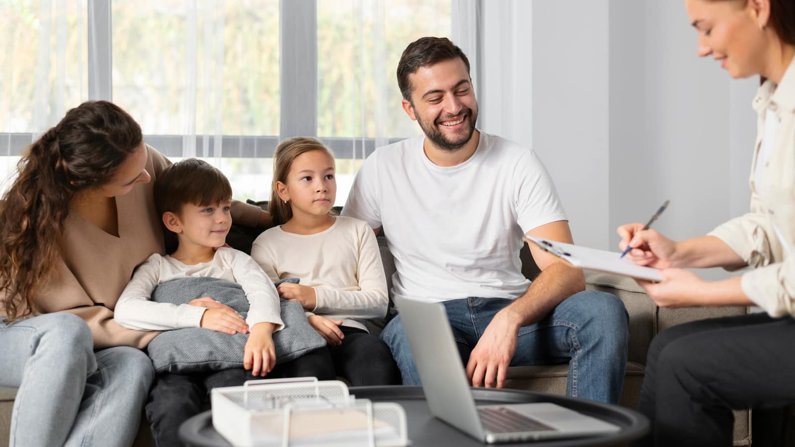 Two parents and their young son and daughter on a sofa in a family therapy session as the therapist writes