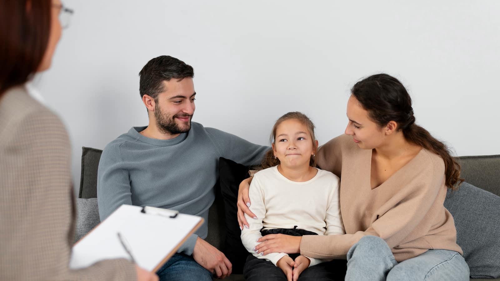 A girl sitting between her mother and father on a sofa, all three looking toward a therapist with a clipboard
