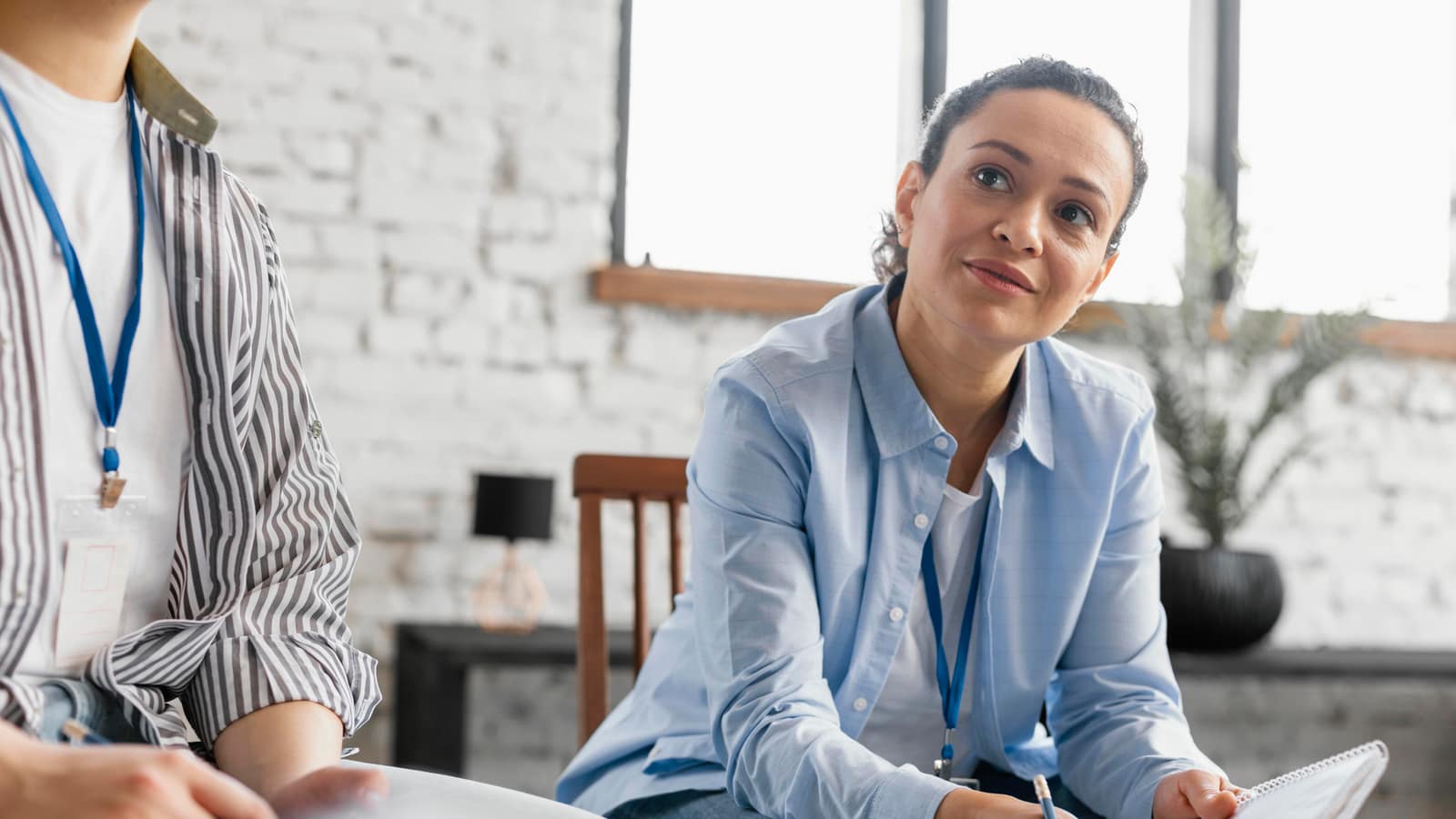 Two clinicians wearing staff lanyards in a bright office, one taking notes on a spiral pad as the other listens