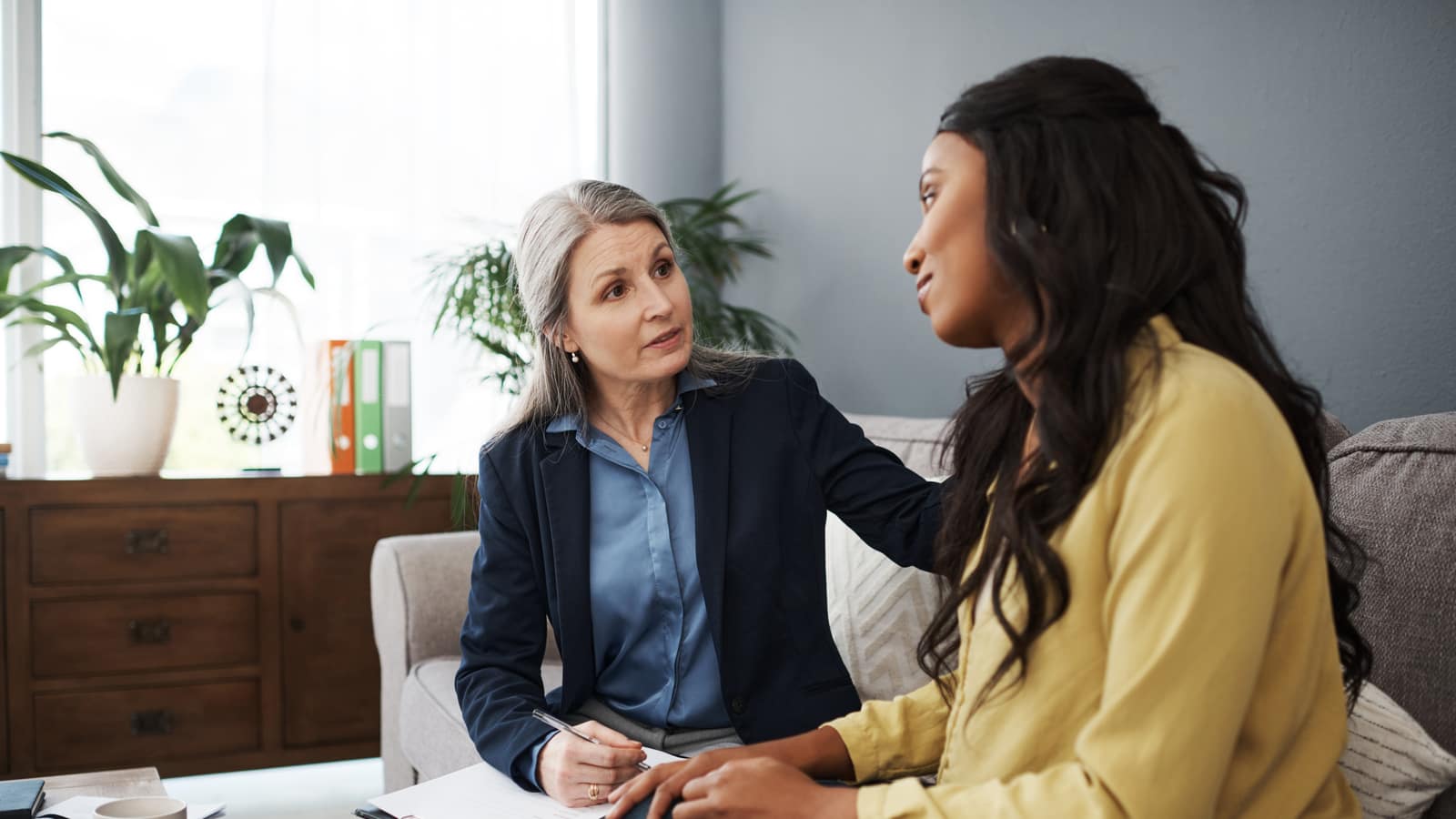 An experienced clinician with a hand on a younger woman’s shoulder, notepad in her lap, in a consulting room