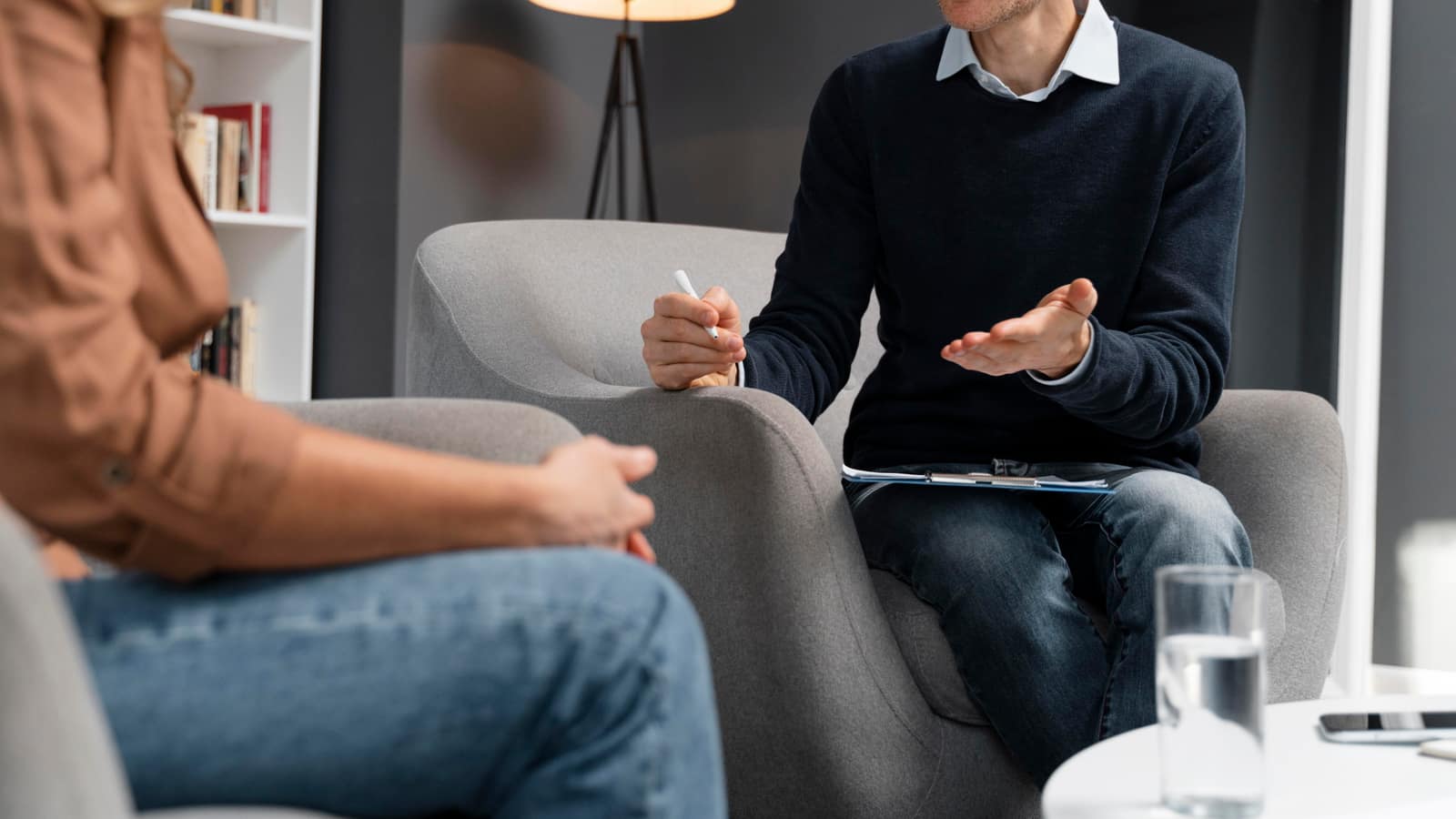 A therapist gesturing with a pen and clipboard on his lap while a client listens from the facing armchair