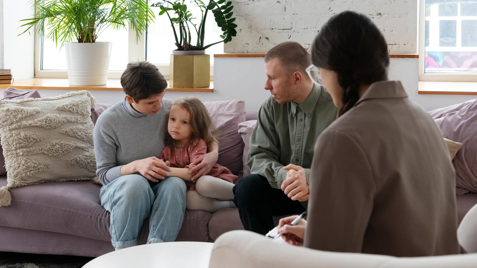 A small girl on her parent’s lap between both parents on a sofa, with a therapist taking notes opposite them