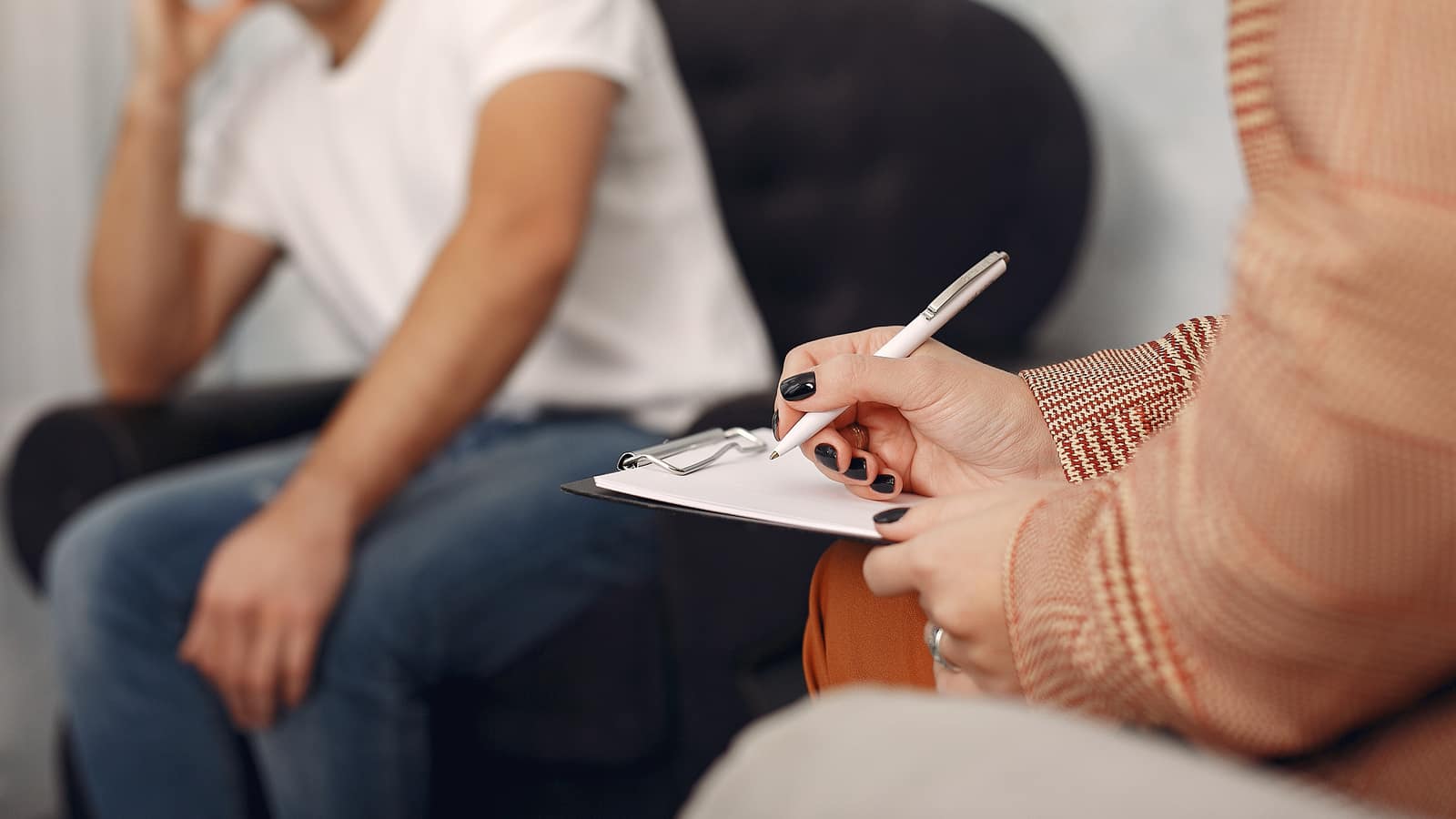 Close view of a therapist writing case notes on a clipboard, with a client out of focus in the background