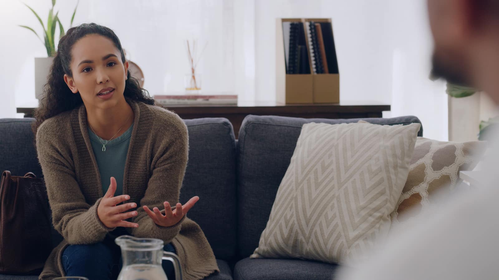 A woman on a sofa speaking and gesturing mid-sentence during a counseling session, the listener in soft focus